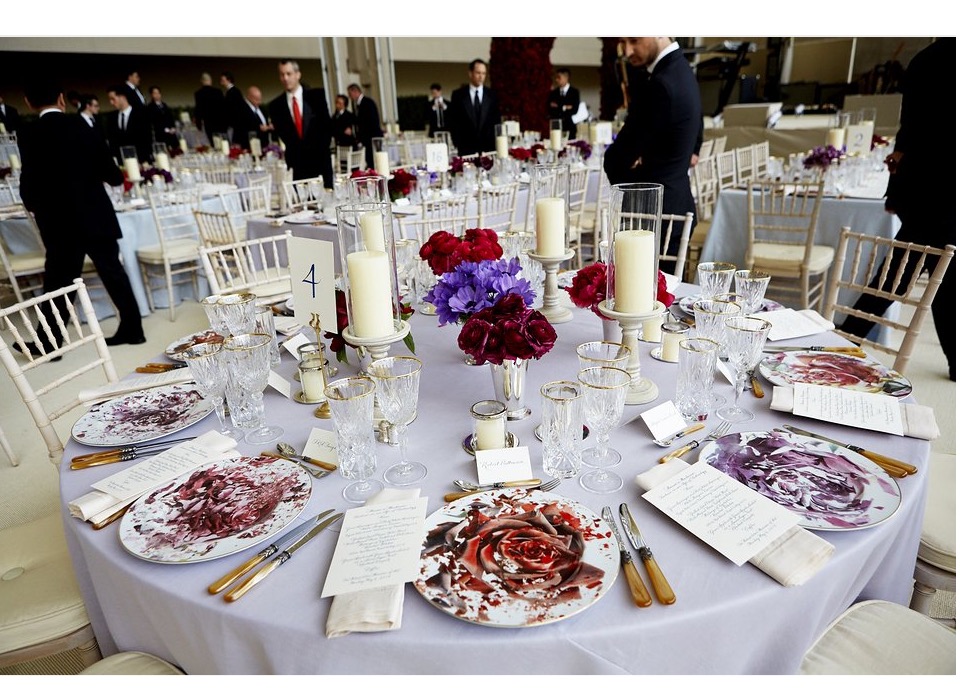 2016 Met Gala Dinner table covered in lavender linen with custom-designed charger plates featuring a modern take on the rose motif. In the center of the table, a grouping of tall pillar candles nestled among red, burgundy, and lavender roses in silver vessels. Image credit Taylor Jewell/Vogue Magazine.